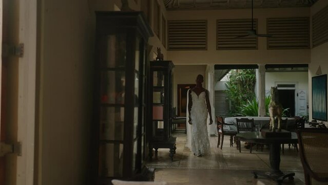 Gay bipoc man walks through luxury retro villa with vintage furniture in white dress with chiffon veil. Black person goes out to swimming pool on paradise island to make wedding photo session.