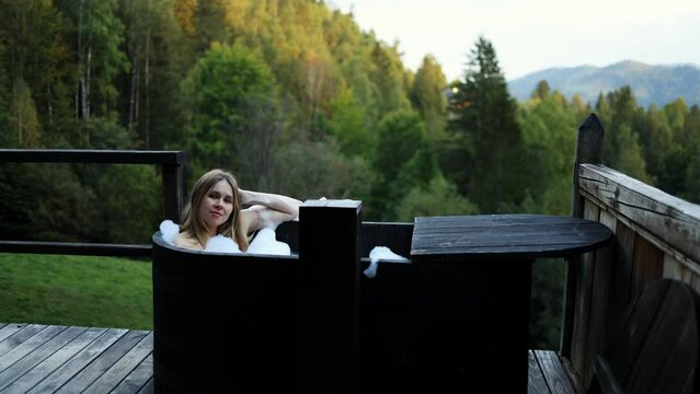 beautiful girl takes a bath in the evening in a wooden black bath outside on the background of mountains and forest.