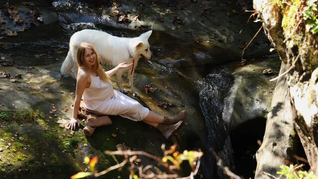 a pregnant girl laughs and sits on a stone with a white shepherd dog in the mountains near the river. top view