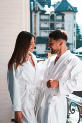 Close up of young couple in bathrobe standing on hotel balcony against mountain background - obrazy, fototapety, plakaty