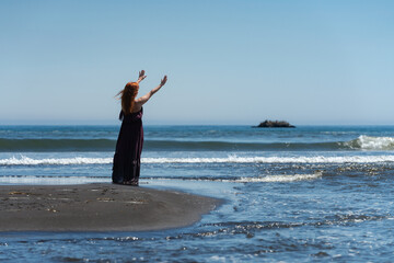 Rear view of happy redhead woman in dark purple long dress standing on summer sandy beach with...