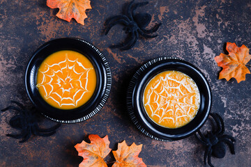 Pumpkin cream soup with spider web of fresh cream in a black bowl with spider decorations. Halloween food concept.