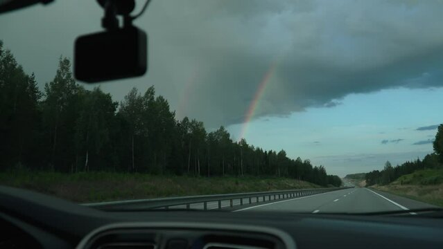View from car window onto highway and rainbow in the sky after rain, handheld shot. Traveling by car, driving along empty country road with dividing fence.