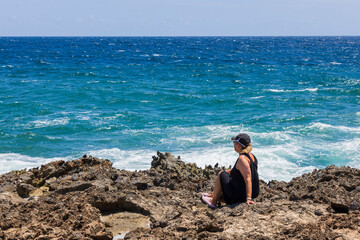 Beautiful view of rocky coast of Caribbean Sea and woman sitting on shore looking into distance. Aruba Island.