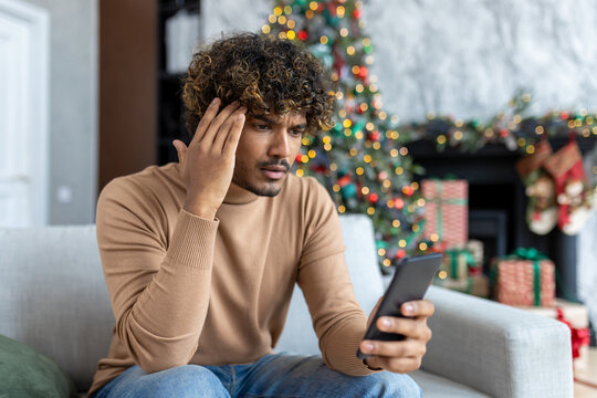Upset sad and disappointed man received message online about bad news, hispanic sitting on sofa in living room, using app on smartphone, depressed unhappy reading internet pages.