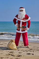 Santa Claus at sea on a hot summer day. Santa Claus stands barefoot on a sandy beach in front of the sea. Bag of gifts on the sand.