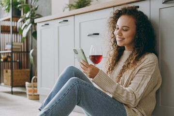 Close up young housewife woman of African American ethnicity wear casual clothes sweater drink wine use mobile cell phone sit near table in light kitchen at home alone. Lifestyle cooking food concept.