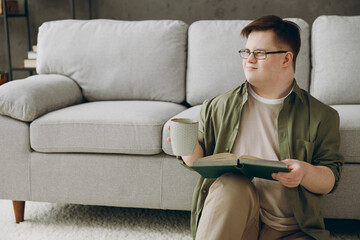 Young smart man with down syndrome wear glasses casual clothes reading book drink tea sit near grey...