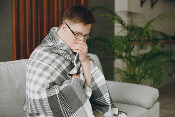 Young sick ill tired exhausted man with down syndrome wears glasses wrapped in plaid sits on grey...