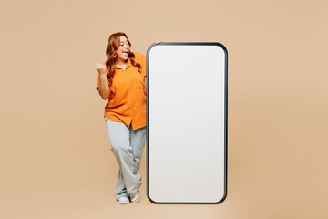 Full body young overjoyed chubby overweight woman wear orange shirt casual clothes big huge blank screen area mobile cell phone doing winner gesture isolated on plain beige background studio portrait.