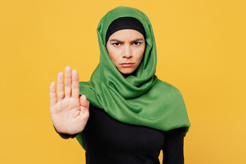 Young serious sad arabian asian muslim woman wear green hijab abaya black clothes showing stop gesture with palm isolated on plain yellow background. People uae middle eastern islam religious concept.