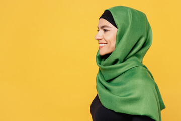 Close up side view young smiling happy cheerful arabian asian muslim woman wear green hijab abaya black clothes isolated on plain yellow background. People uae middle eastern islam religious concept.