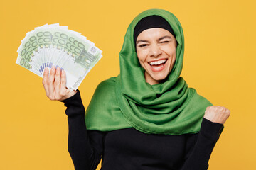 Young winner arabian asian muslim woman wearing green hijab black clothes hold fan cash money in dollar banknotes isolated on plain yellow background People uae middle eastern islam religious concept
