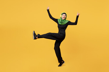 Fototapeta na wymiar Full body young asian muslim woman wear green hijab abaya black clothes listen to music in headphones raise up leg hands dance isolated on plain yellow background. People uae islam religious concept.