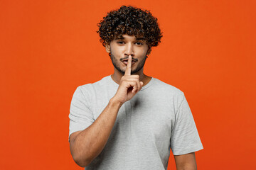 Young secret smiling fun happy Indian man he wears t-shirt casual clothes say hush be quiet with finger on lips shhh gesture isolated on orange red color background studio portrait. Lifestyle concept.