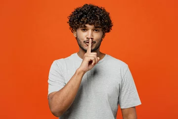 Fotobehang Young secret smiling fun happy Indian man he wears t-shirt casual clothes say hush be quiet with finger on lips shhh gesture isolated on orange red color background studio portrait. Lifestyle concept. © ViDi Studio