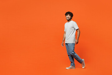 Fototapeta na wymiar Full body side profile view young smiling happy Indian man he wearing t-shirt casual clothes walk going looking camera isolated on orange red color wall background studio portrait. Lifestyle concept.