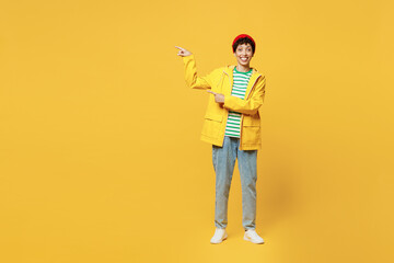 Fototapeta na wymiar Full body young woman wearing waterproof raincoat outerwear red hat point finger aside on area isolated on plain yellow background studio portrait. Outdoors lifestyle wet fall weather season concept.