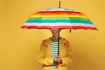 Young smiling woman wearing waterproof raincoat outerwear hold in hand cover face with striped umbrella isolated on plain yellow background studio. Outdoors lifestyle wet fall weather season concept. - obrazy, fototapety, plakaty
