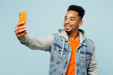 Young man of African American ethnicity wear denim jacket orange t-shirt doing selfie shot on mobile cell phone post photo on social network isolated on plain pastel light blue cyan background studio.