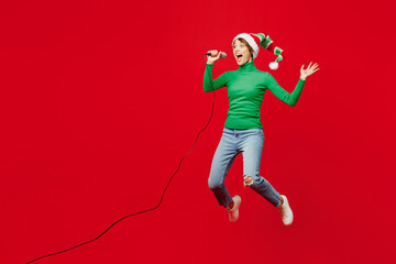 Full body young singer fun woman wear green turtleneck Santa hat posing jump high sing song in microphone isolated on plain red background. Happy New Year 2024 celebration Christmas holiday concept.