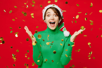 Young woman wear warm green turtleneck Santa hat posing spread hands look camera in falling confetti around isolated on plain red background. Happy New Year 2024 celebration Christmas holiday concept.