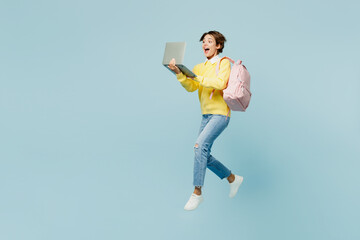Full body young woman IT student wear casual clothes sweater hold backpack bag jump high hold use work on laptop pc computer isolated on plain blue background. High school university college concept.