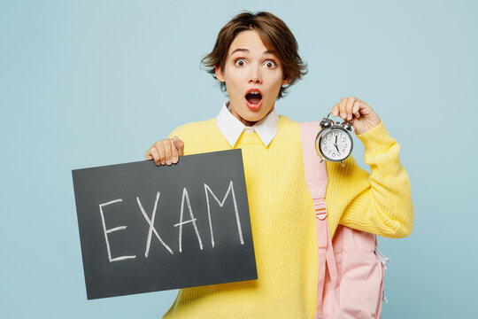 Young woman student wear casual clothes yellow sweater backpack bag hold card sign with exam title text alarm clock being late isolated on plain blue background High school university college concept