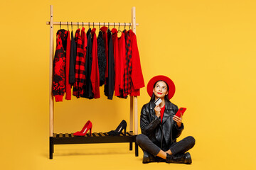 Full body young minded pensive woman wear red hat sit near rack of casual clothes use mobile cell...
