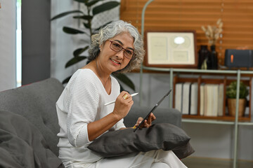 Senior Asian woman using a digital tablet while sitting on the sofa, looking at camera