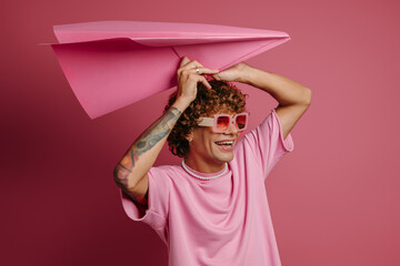 Happy young curly hair man holding big paper airplane upon head while standing on pink background
