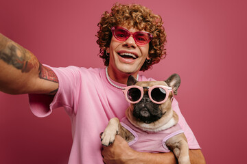 Joyful young man holding French Bulldog pet in funky glasses and making selfie on pink background