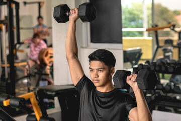 A young asian man looking focused while doing a set of seated alternate dumbbell presses at the gym. Shoulder workout and training.