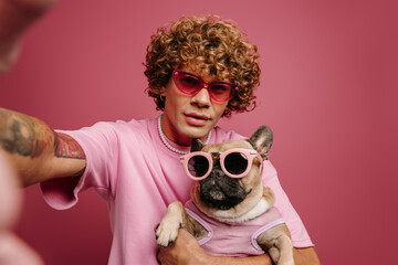 Fashionable young man holding French Bulldog pet in funky glasses and making selfie on pink background - 661888253