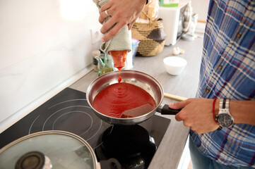 View from above of a male chef adding a fresh passata from ripe organic tomatoes into a frying pan, while cooking a delicious meal in the home kitchen. Man cooking at modern minimalist home kitchen