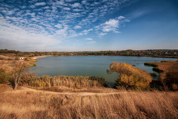 Autumn landscape on the river bank with a single tree with yellow leaves, water and dry grass around it and beautiful clouds in the blue sky, a long path is visible near the pond 
