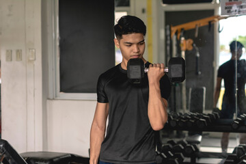 A young serious asian man doing standing alternate bicep curls. Working out and training arms at the gym.