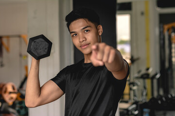 A young handsome asian man pointing to the camera while doing standing alternate bicep curls....