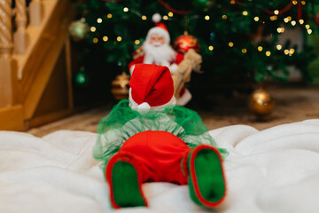 A newborn baby in a Christmas elf costume lies on his stomach on a white blanket near a Christmas...