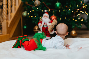 A little newborn baby in Christmas elf clothes is lying on stomach on a white blanket and looking at Christmas tree with bokeh at Christmas. No face. Back view. 