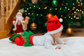 A newborn baby in a Christmas elf costume lies on his stomach on a white blanket near a Christmas...