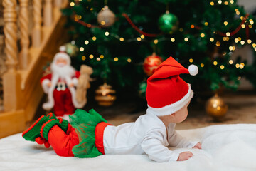 A newborn baby in a Christmas elf costume lies on his stomach on a white blanket near a Christmas tree with bokeh for Christmas. Back view. 
