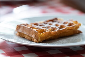 A portrait of a belgian tasty delicious waffle with powder sugar as a topping on it, lying on a white plate on a red table cloth ready to be eaten during breakfast or lunch or as a snack.