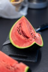 Vertical portrait of a quarter slice of a green cut watermelon lying on a plastic cutting board next to a knife. The piece of the fruit is ready to be eaten and you can see black seeds in the red pulp