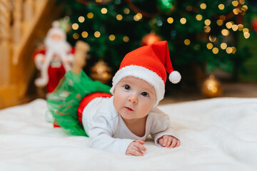 Baby girl with Christmas elf clothes is lying on a white blanket near Christmas tree with bokeh. ...