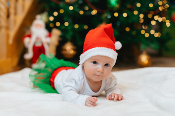 Baby girl with Christmas elf clothes is lying on a white blanket near Christmas tree with bokeh.  Christmas concept. Infant baby in her first Christmas. Funny baby. 