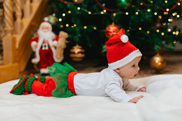 Baby girl with Christmas elf clothes is lying on a white blanket and showing hr tongue near...