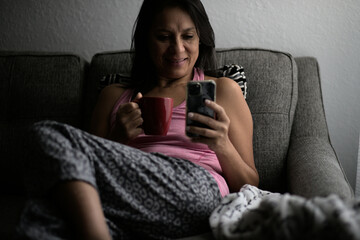 Smiley mature woman relaxing with a cup of coffee looking at phone early in the morning, waking up in pajamas.