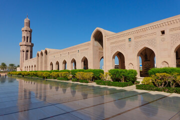 Fototapeta premium Exterior of the colonnade with the minaret of the Grand Mosque of Sultan Qaboos composed of many arches reflected on marble adorned with hedges and blue sky. Oman.