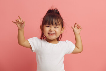 Joyful three years old Asian ethnicity little girl raises her hands up, expresses a cheerful mood, smiles happily standing on a pink isolated.